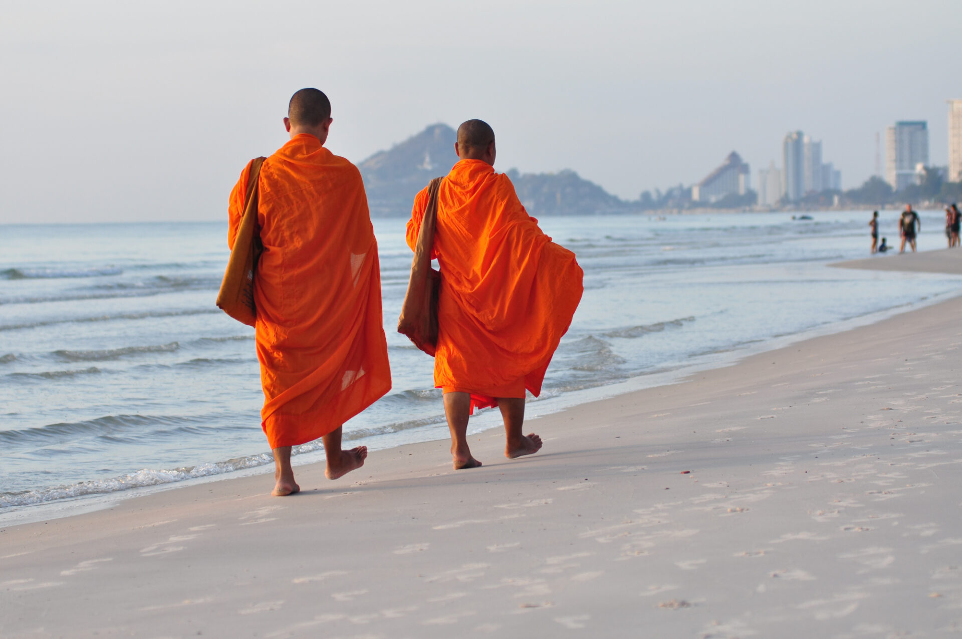 To buddhistmunker i oransje kapper går barbeint langs stranden i Hua Hin, med bylivet og åsene i bakgrunnen.